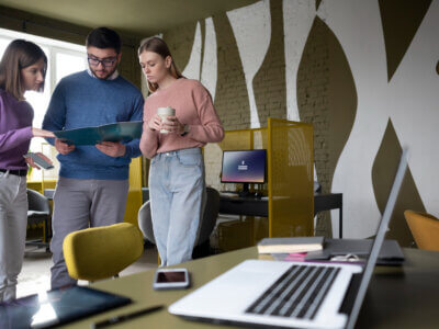 Three people stand in a modern office in Israel, reviewing documents together. Two laptops, a tablet, and a phone are visible on a desk in the foreground, hinting at the role of AI in todays jobs. Bright light comes in through large windows.
